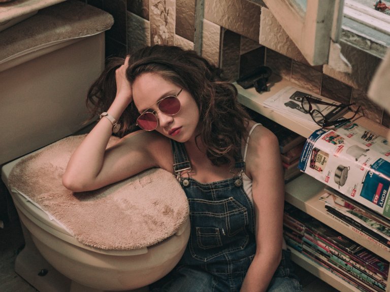 A frustrated woman sitting on the floor of a bathroom, resting her arm on the carpeted lid of a toilet.