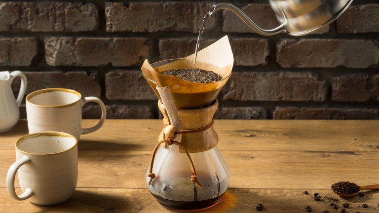 Gooseneck kettle pouring water into pour-over coffee carafe on table next to coffee beans and mugs
